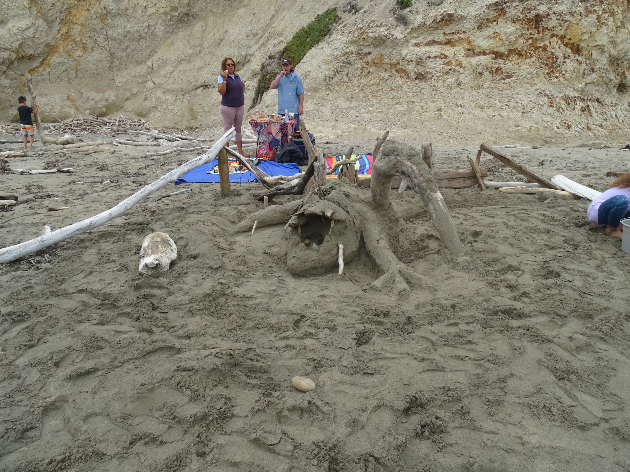 A sand sculpture of a sea monster with a large open mouth and irregular legs made of sand and driftwood.
