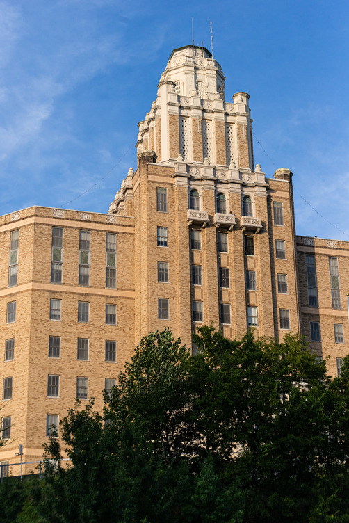 A large tan brick building. Over seven stories tall behind green trees 