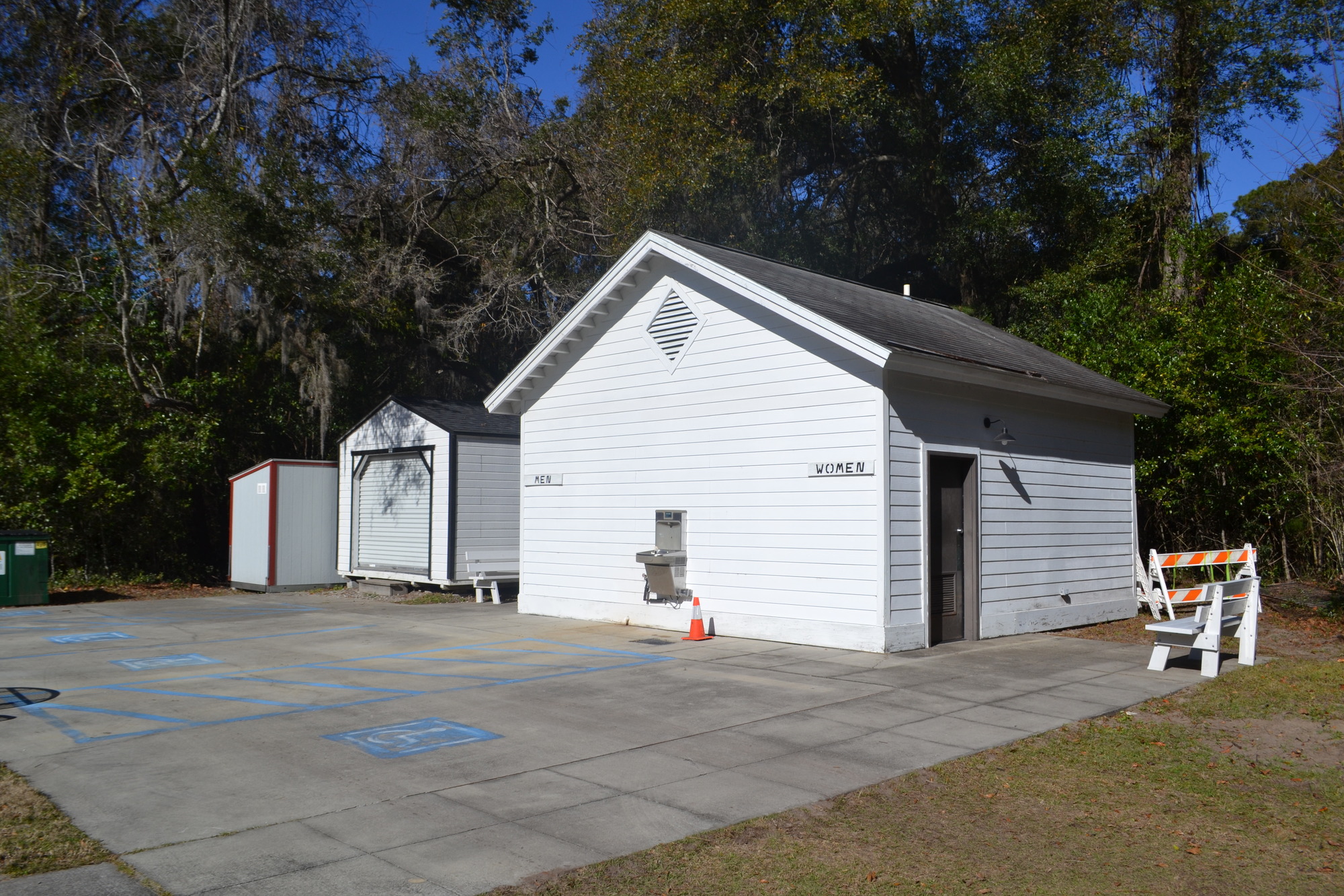 A small white building with signs on the left and right side that say “Men” on the left and “Women” on the right. It sits on a paved accessible parking lot. Doors to each restroom are on either side of the building. A water fountain and water bottle station are on the center of the front wall.
