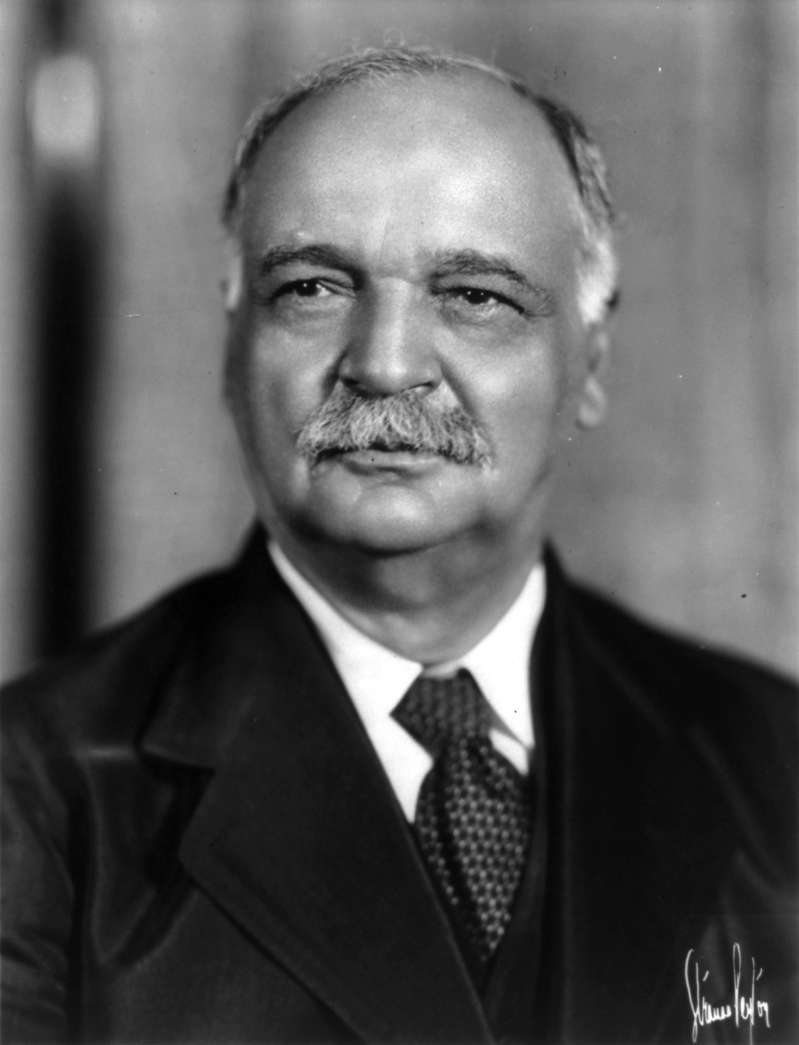 A soft focus 1928 photo captures the bust portrait of a gray haired and mustached man in a suit. 