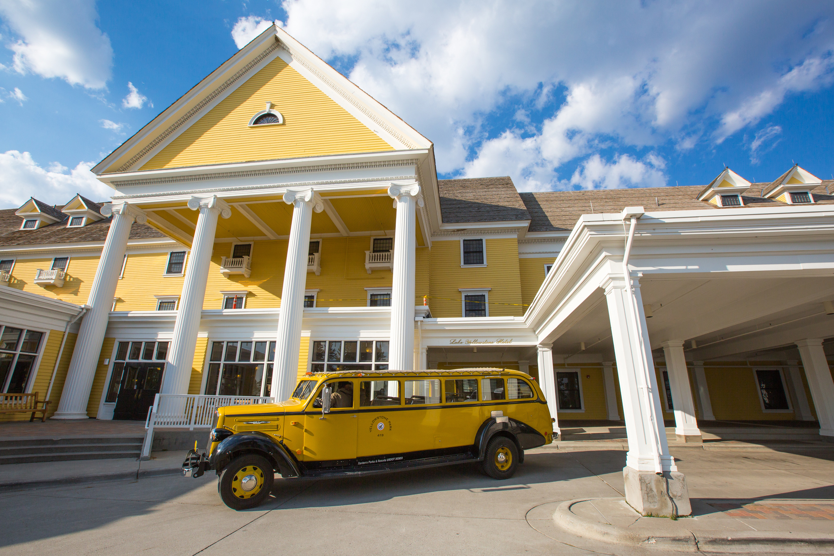 A 1930s style yellow touring bus sits in front of a colonial styled hotel which is yellow with 4 white pillars in front.