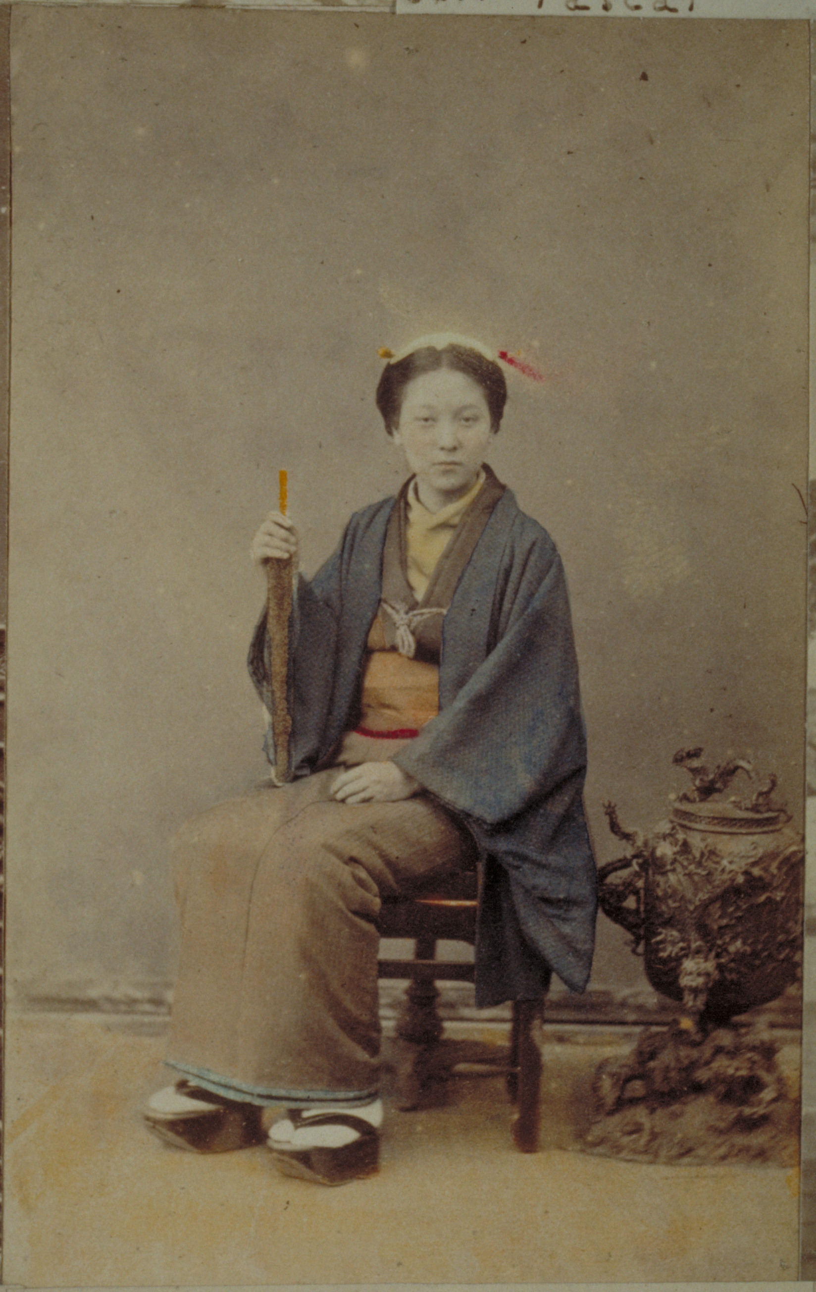 Black-and-white photograph with colored-in blue, yellow, orange and red details representing a Japanese woman sitting on a chair by an urn and holding an unfolded umbrella. She is photographed against a plain wall background.