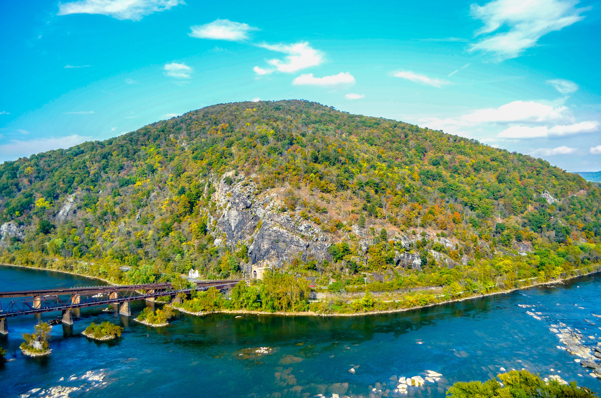 View of Maryland Heights from the Split Rock Overlook along the Loudoun Heights Trail. The image has very bright colors because of the saturation and the trees are various shades of green. You can see the rock face of Maryland Heights and the train tracks and pedestrian bridge coming out of the Maryland side of the mountain. 