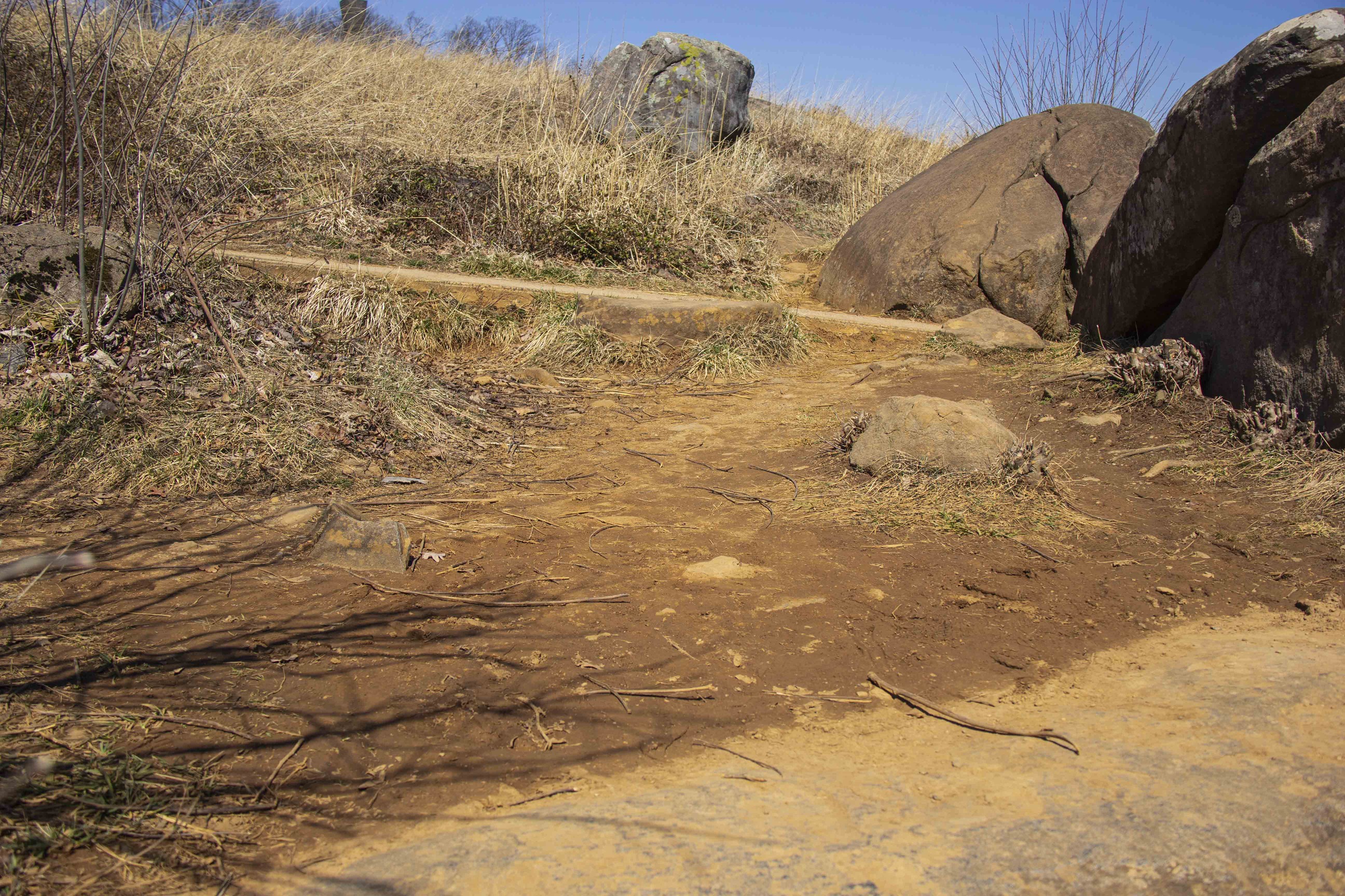 This is a view of unhealthy and eroded soil. The center of the frame is full of dark brown earth with small rocks little plant life. Ahead is a dirt path leading to two large boulders on the right. Past the path is tall dark grass in contrast to the barren region in the center.