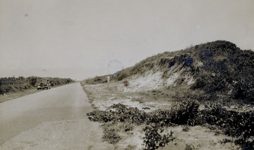 Black and White photo.  Car parked.  Person standing on road side.