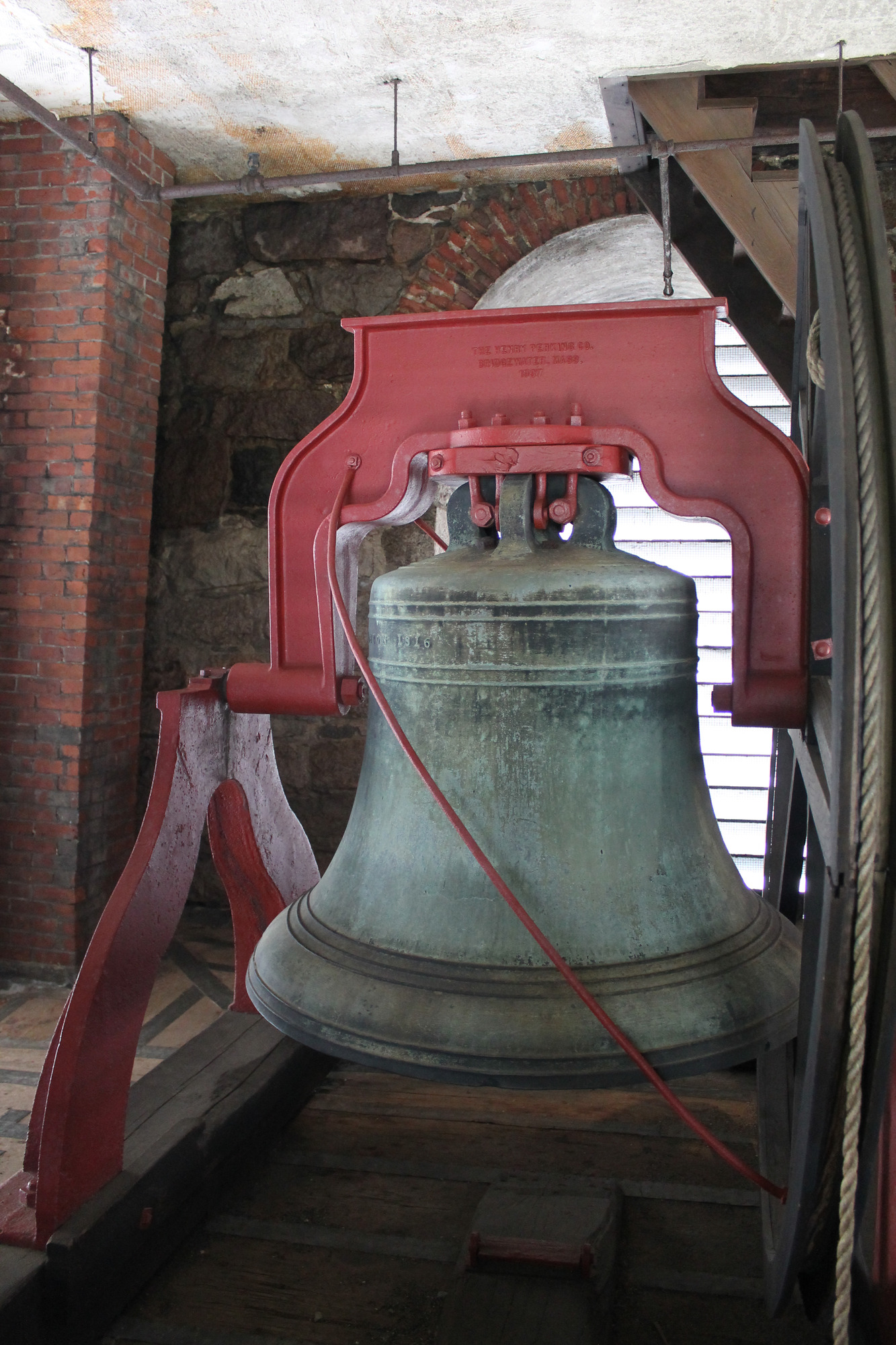 An interior image of the bell of King's Chapel. The bell is a variety of colors including black, grey, green, and brown. It is a large bell that is held up by a red, metal system. The bell is located in the tower of the chapel which is made of brick and wood.