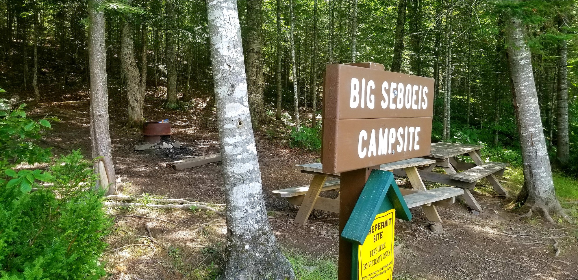 A brown sign with white text reads "Big Seboeis Campsite" with picnic tables in the background.