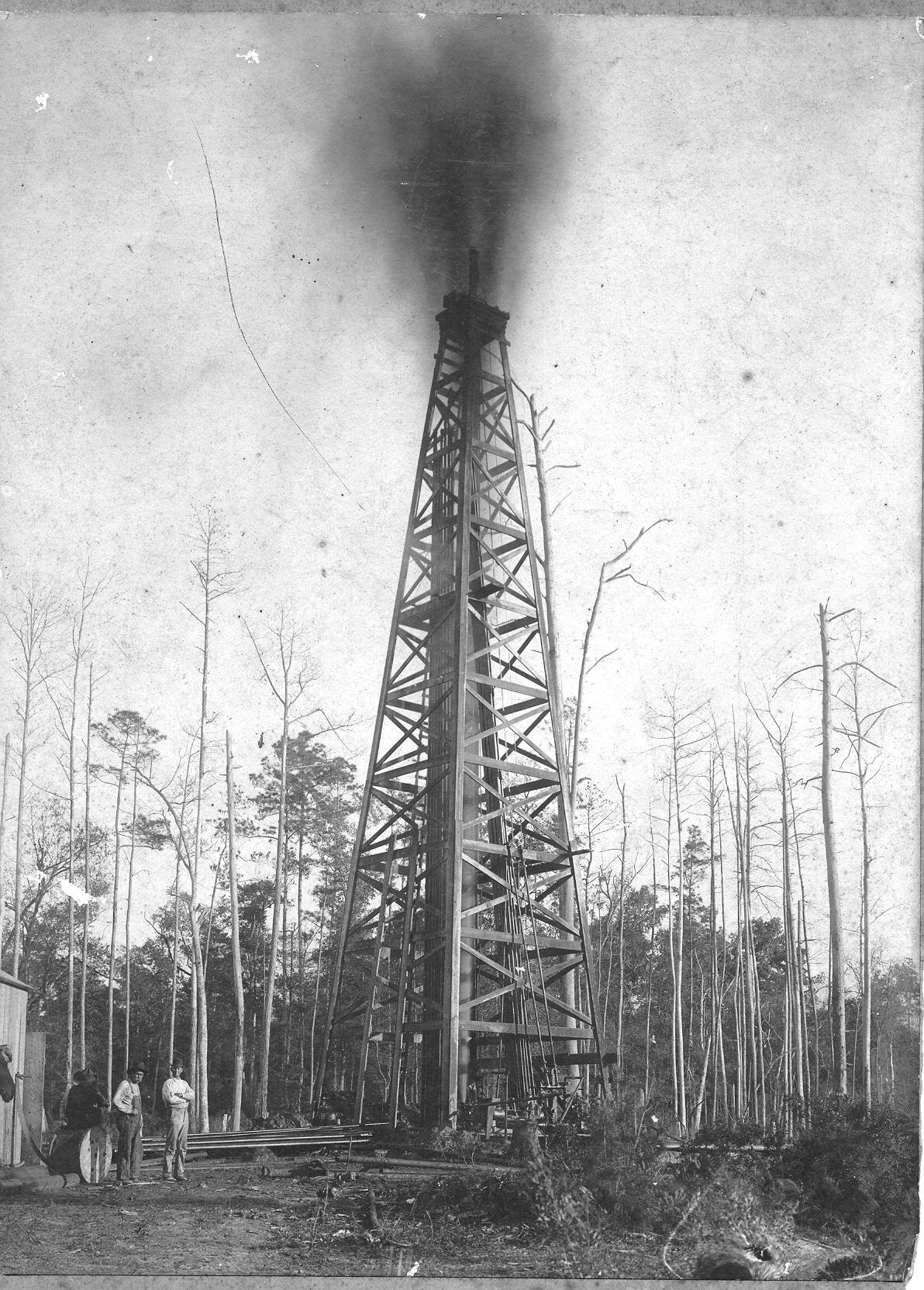 Historic black and white photo of a tall wooden oil derrick gushing oil into the sky while a few people stand near its base.