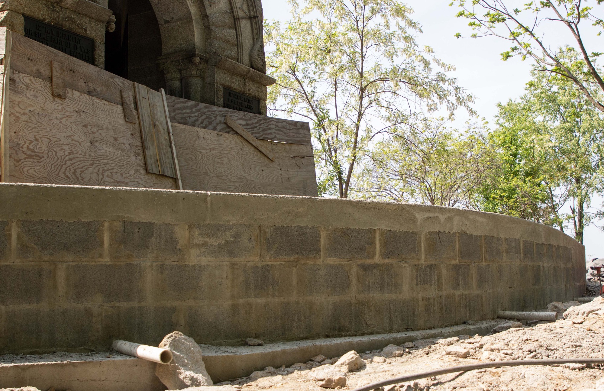 A cinderblock retaining wall with a concrete sidewalk stands elevated along a large stone monument, drainage pipes and rocks are at the base of the wall.