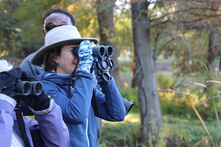 A woman wearing a wide-brimmed hat, blue jacket, and patterned gloves holds a pair of binoculars up to her eyes, looking towards the right side of the frame. Her mouth is slightly open in a smile. Behind her, a man with dark skin and sunglasses is partially visible, also looking in the same direction. In the foreground to the left, another person's arm is visible, holding a pair of binoculars. The background is a blurred outdoor scene with trees and some dry grasses, suggesting an autumn or late fall setting.