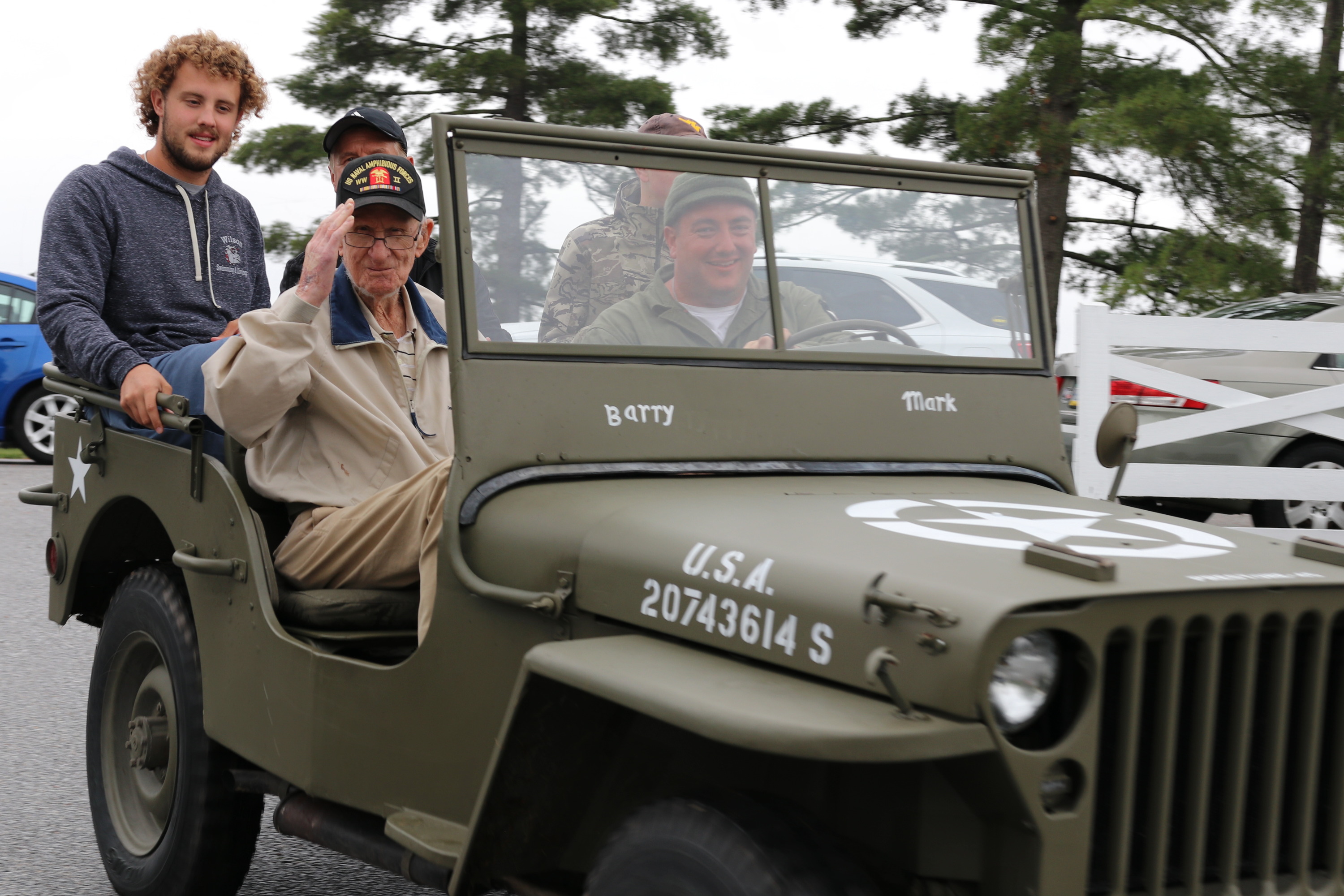 A man in a ballcap and jacket sits in the front seat of a Jeep, saluting the camera, with others seated behind him. 