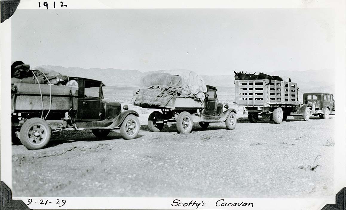 This is an historic black and white photograph from the Scotty's Castle Historic Photograph Collection, Death Valley National Park of four vehicles in a line stopped in desert. First vehicle is a sedan. Second vehicle is a livestock truck filled with horses and mules. Third truck piled above cab with canvas covered supplies. Final truck has canvas covered supplies. Inscriptions in black ink along upper and lower border.