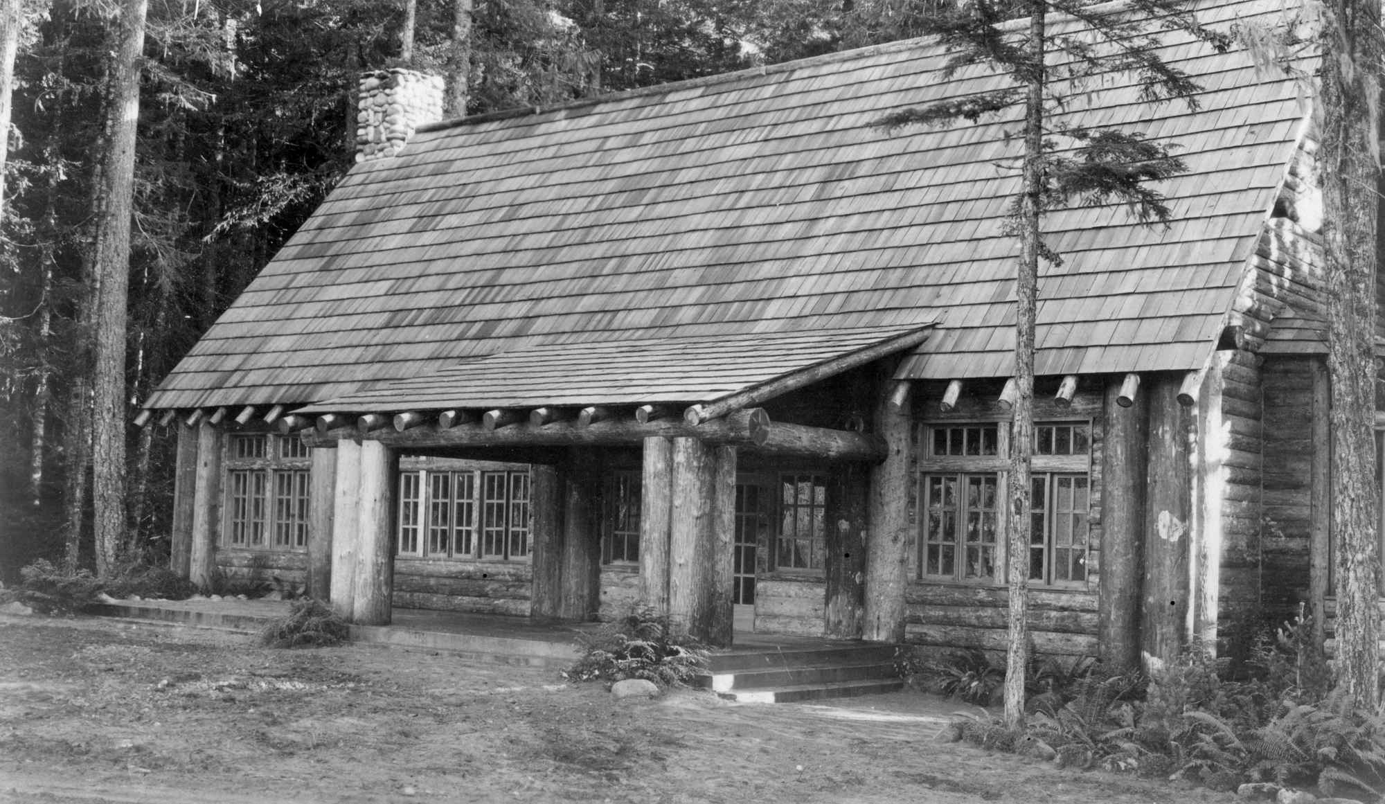 Black and white historic photo of a large log building in a forest with a steep shingle roof and a porch supported by log beams. Ferns ad plants grow around the edge of the building and porch. 