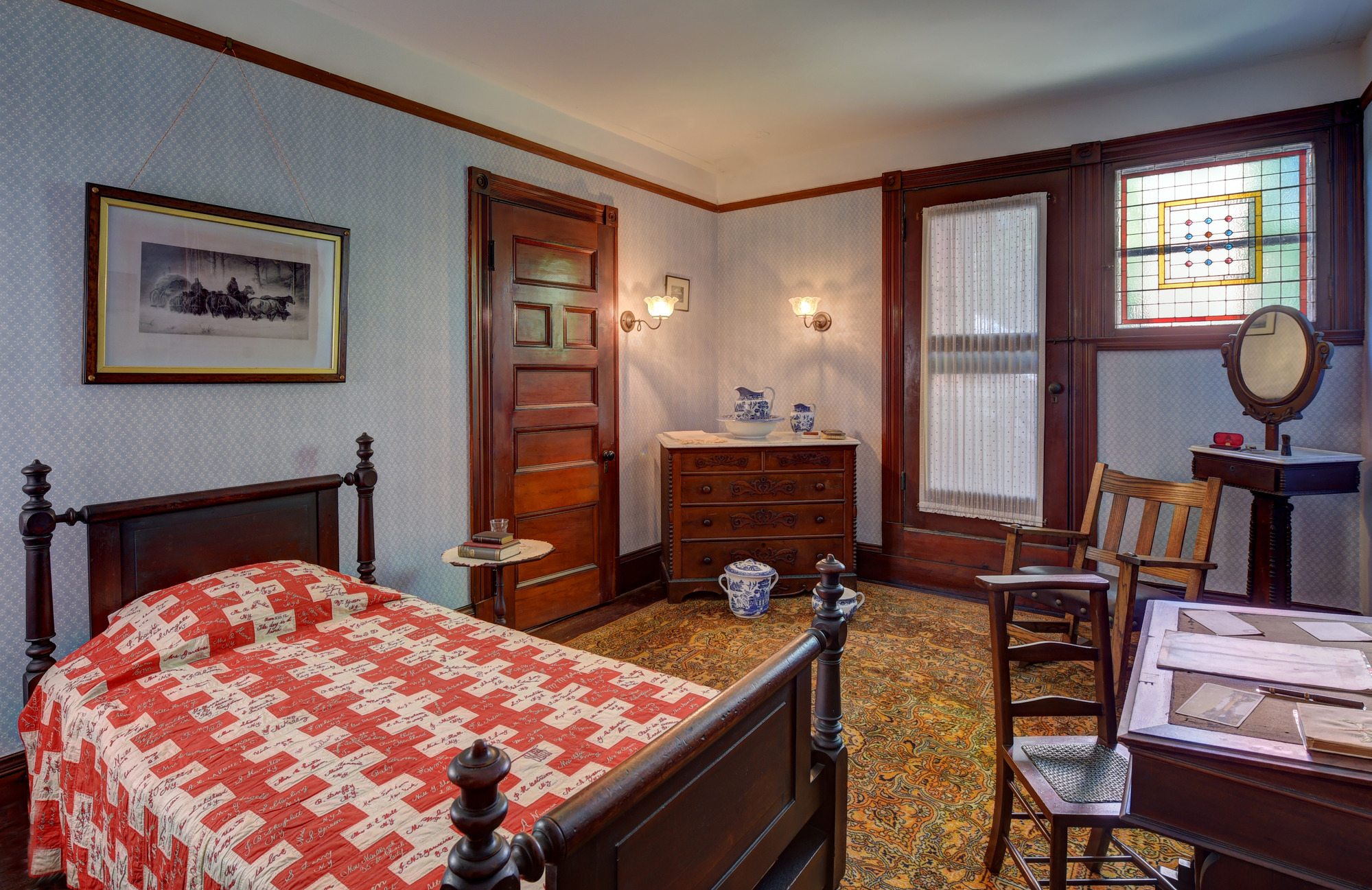 A small blue and white wallpapered bedroom with a single bed, desk, dresser, and shaving stand. There is a red and white checkered quilt on the bed. There is a yellow, orange, green, and pink rug on the floor. 