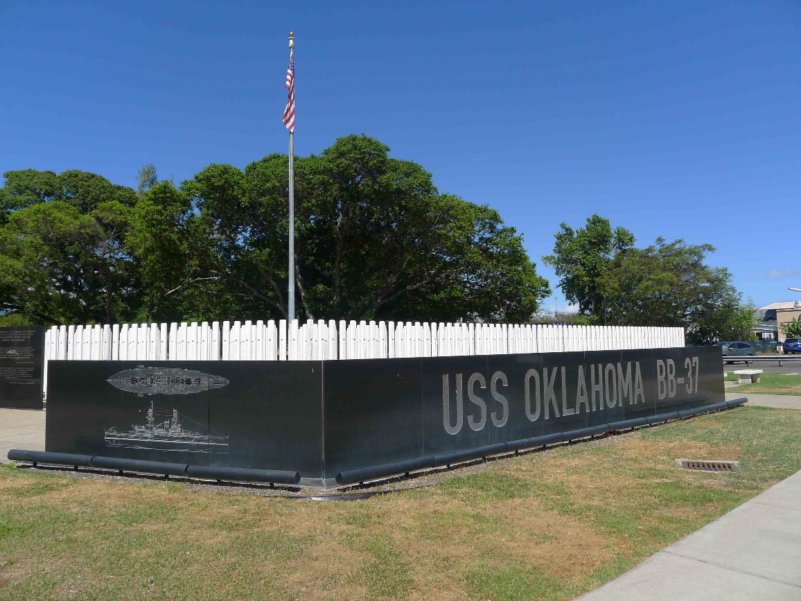 The USS Oklahoma Memorial is composed of white marble columns and black granite slabs. An American flag stands on a flagpole, and it is surrounded by short turf. 