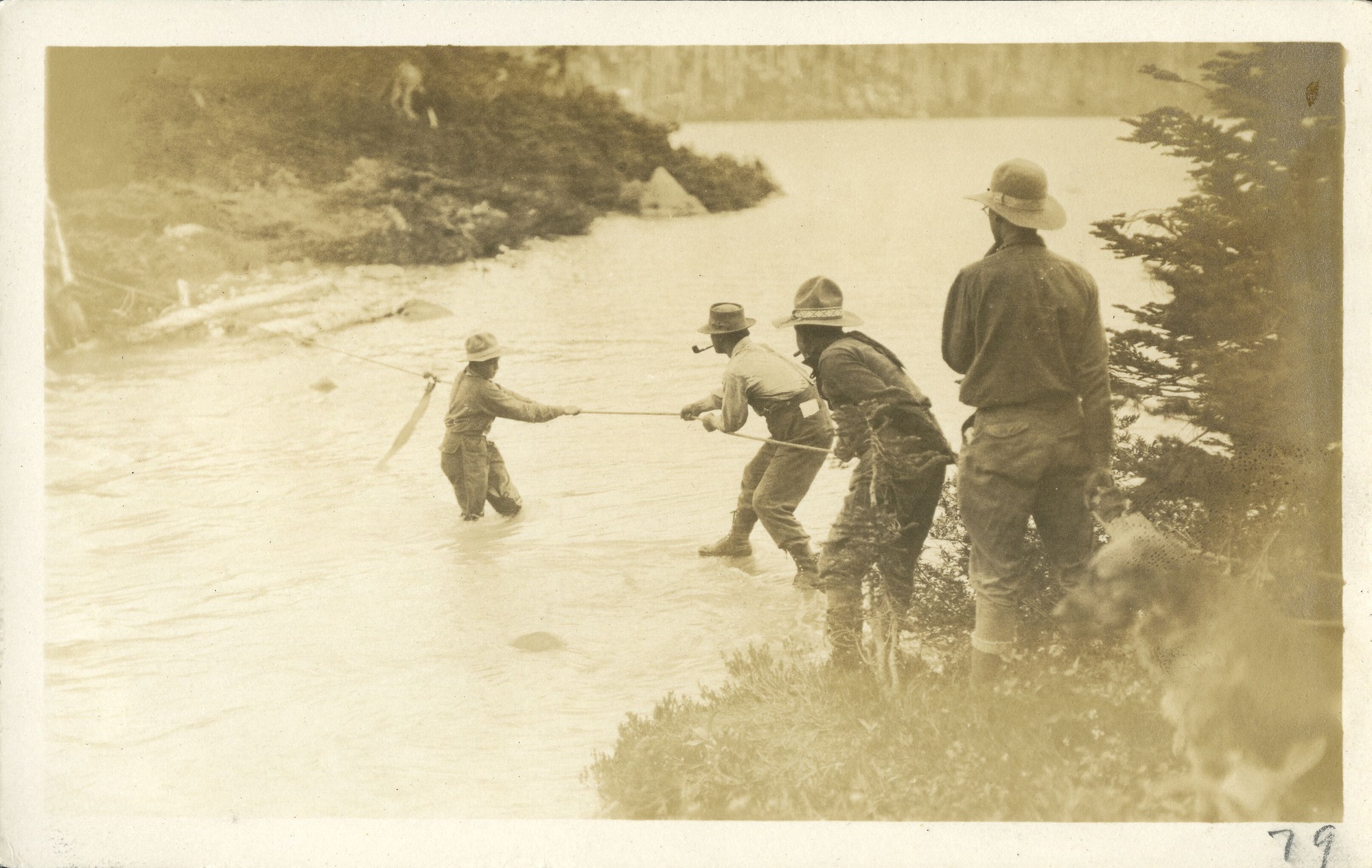 Four men and a dog. One man fording a river with help of rope strung across.