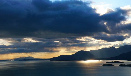 Sunlight shines behind dark clouds and reflects on the turquoise blue of Lake Clark. 