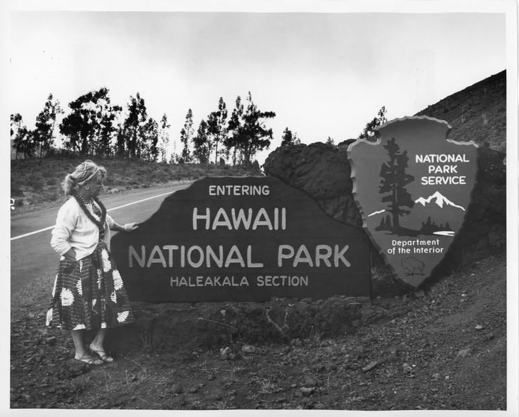 A black and white image of the entrance sign in front of the Haleakalā Section of Hawaii National Park. To the left of the picture is a woman standing next to the sign and posing in front of it. The woman is wearing a white sweater with a floral dress. She also has her hair up with a headband and clips. Around her neck is a necklace and a fabric lei. She is wearing strappy flat sandals. She also has her right hand in the pocket of her dress and has her left hand resting on the entrance sign. The sign is located on the right side of the road. The sign reads, “Entering Hawaii National Park, Haleakala Section.” The sign is also sitting on a rock wall. To the right of the entrance sign is the National Park Service Arrowhead logo with a boulder behind it. The ground that the sign is on is rocky and made of dirt. To the far left of the image is the road that leads into the park. Along the left side of the road there is grass and trees in the distance.