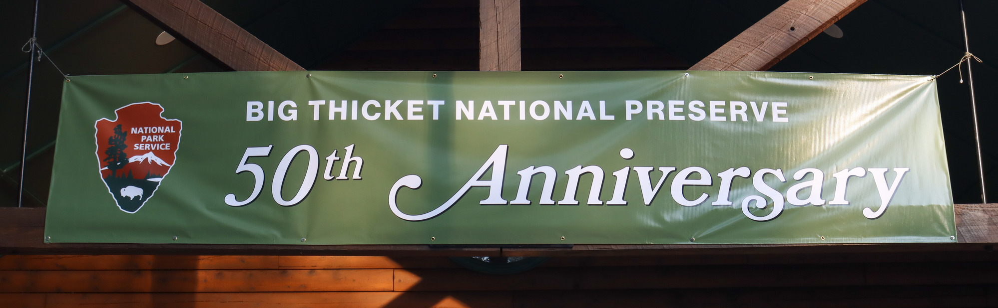 A green banner hanging on wooden beams outside the visitor center. The banner features the National Park Service arrowhead logo and text: Big Thicket National Preserve 50th Anniversary.