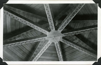This is an historic black and white photograph from the Scotty's Castle Historic Photograph Collection, Death Valley National Park looking up into intricately decorated, open, octagon wood beam ceiling. In middle is circular base of carved beam with wiring for lighting. Radiating out from center are eight square beams with each face carved in same motif. Above are eight more beams with floral motifs tight to wood panel ceiling.