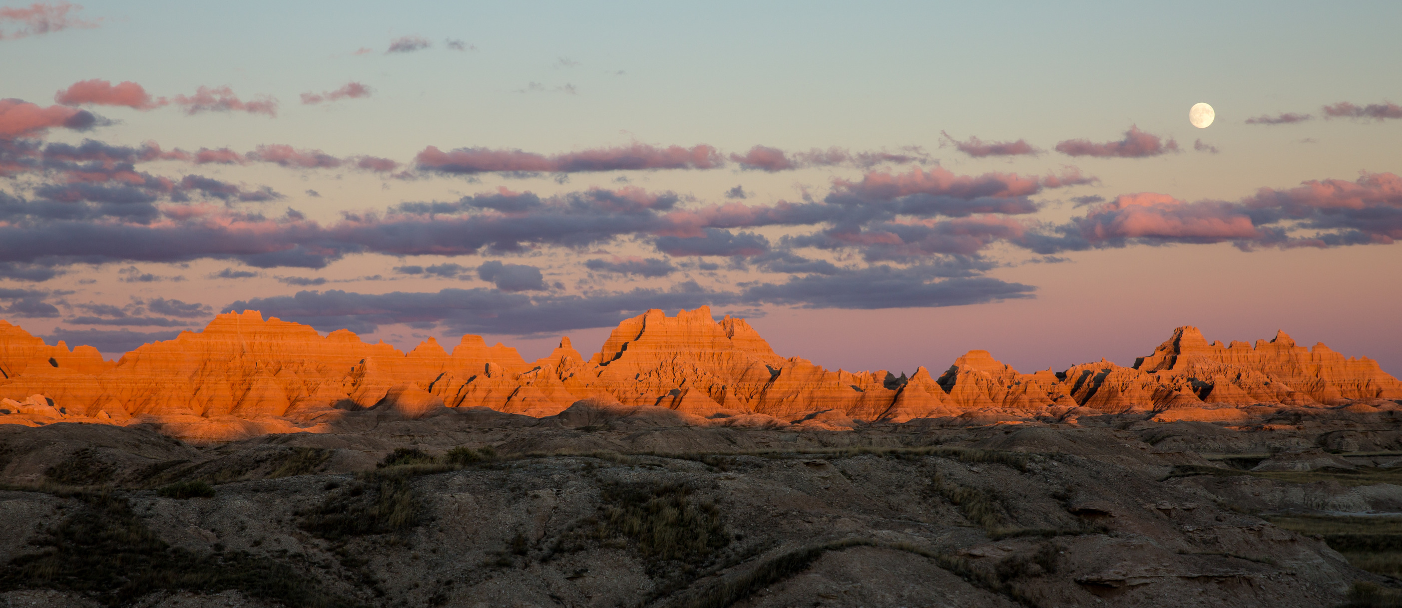 The setting sun illuminates several ridge lines and hills at Badlands National Park. The moon rises in the distance from behind some small clouds. 