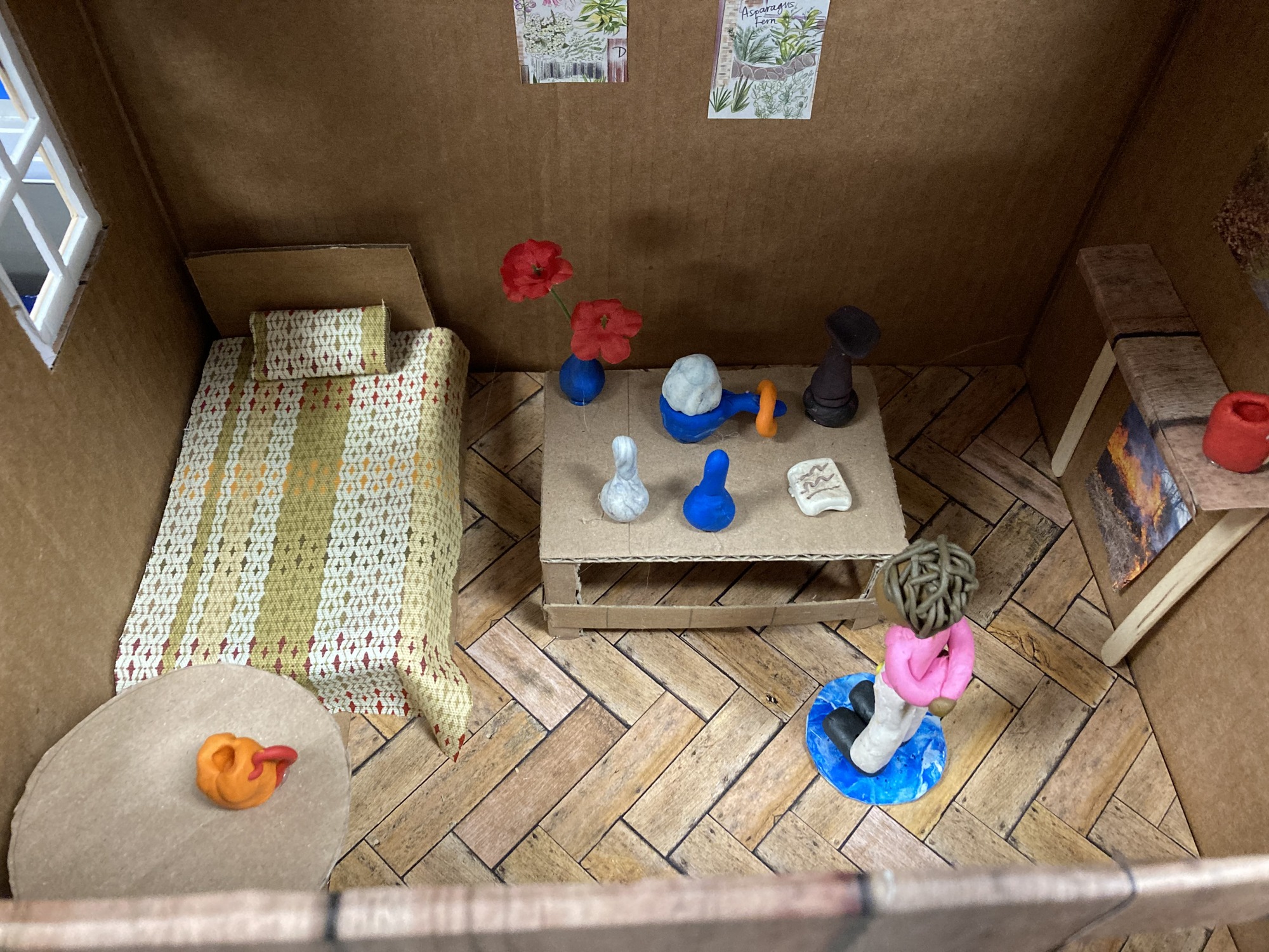 Image of a shadow box of a bed, table with objects, and a person standing on a blue rug. 
