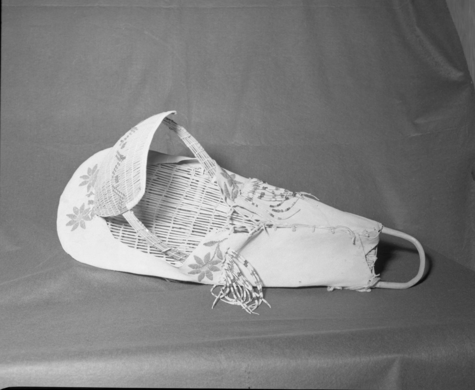 Mrs. Donald Segerstrom Collection. Object: Paiute cradle basket. Dimensions: 34" length. Materials: Willow, cloth, glass beads, buckskin. Technology: Backboard plain twined, up to right slant of turns, hood diagonally twined up to right slant of turns, beadwork of petal and leaves in modified lazy stitch, bead fringe includes "Basket" beads and cornaline d'aleppo beads that may have been excavated. Flowers and petals are worked in a peculiar faceted bead. Photographed by Michael Dixon. Catalogued by Craig Bates and Barbara Beroza. Yosemite Collections, National Park Service. 1/83.