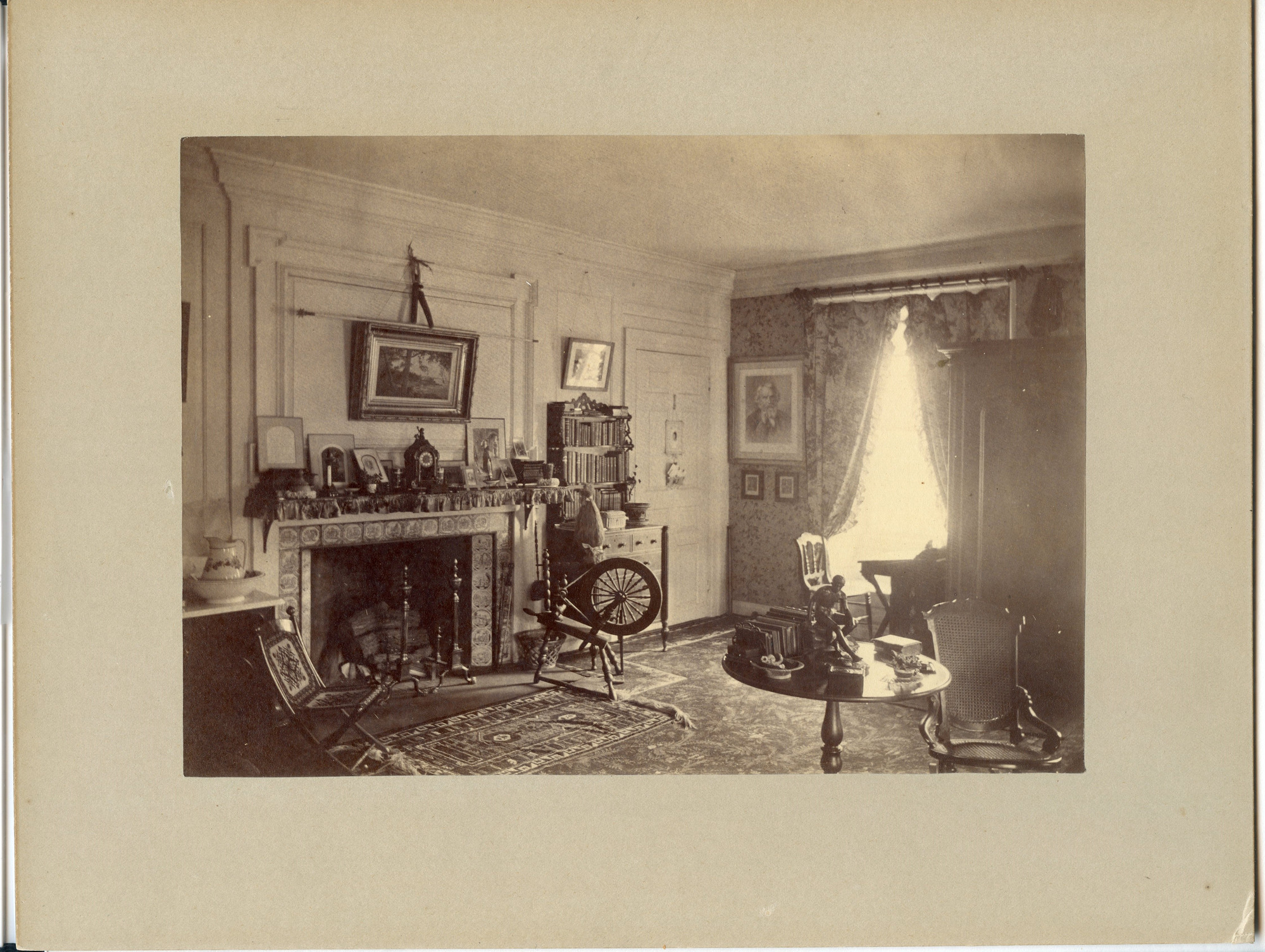 Black and white photograph of Victorian bedroom with fireplace, spinning wheel and bookshelf featured.