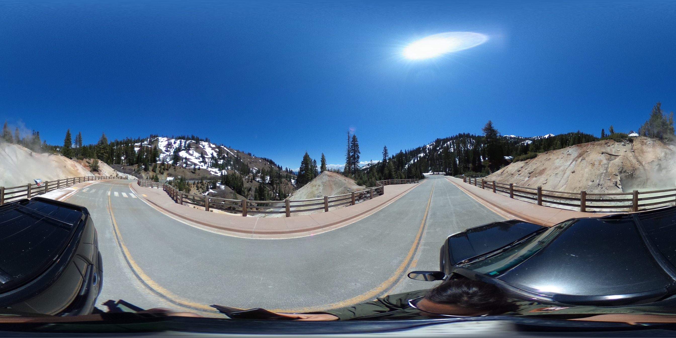 A 360-degree photo of a road passing through an area where steam rises from barren soil. Mountains with patch snow rise in the mid-ground. 