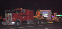 Red semi cab pulling open trailer with strings of lights, people, arrowhead, and slide show on a screen at night.