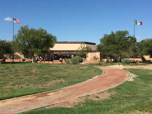 A distant view of sand-colored building with trees, lawn, and red gravel path. Two flags, US on the left and Mexican on the right, fly straight out from their poles.