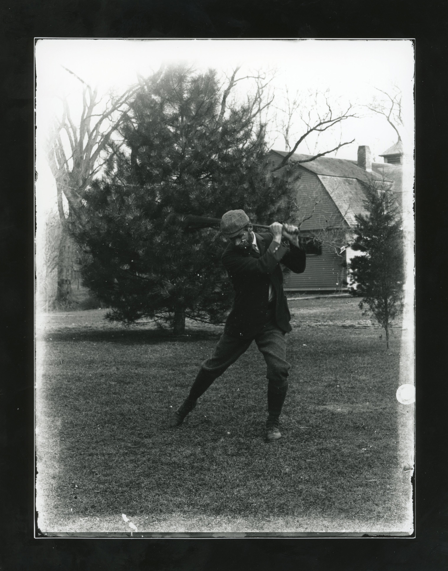 White man in tweed cap at top of golf club swing. Tree and house in background.