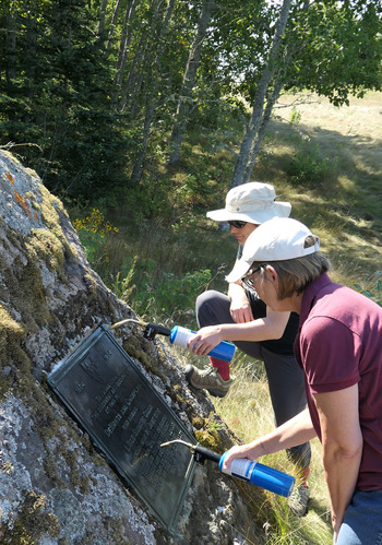 Two people using small propane torches to heat a bronze plaque set into a large boulder .