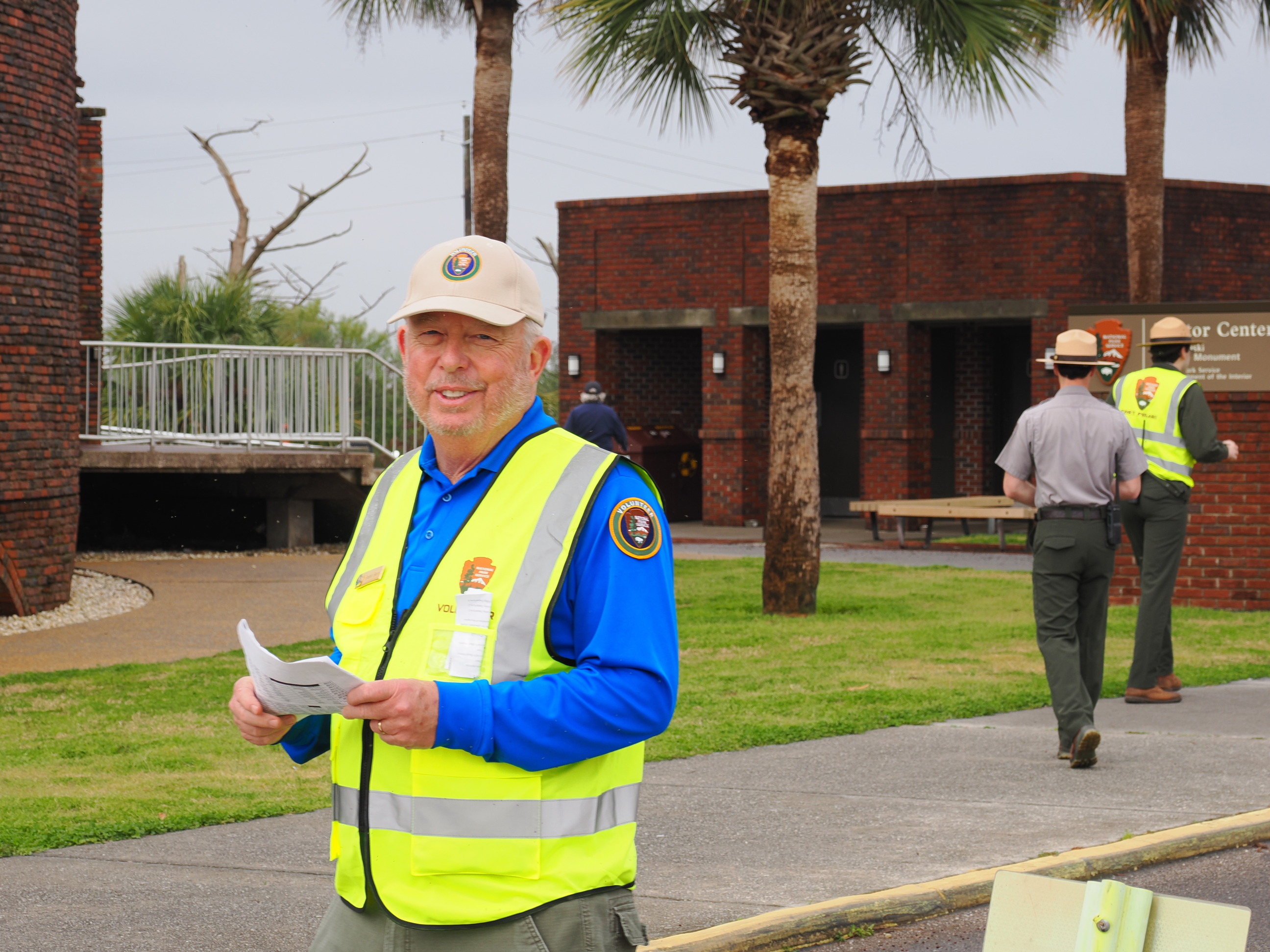 A man in a yellow vest and blue shirt holds a piece of white paper in front of a brick visitor center.