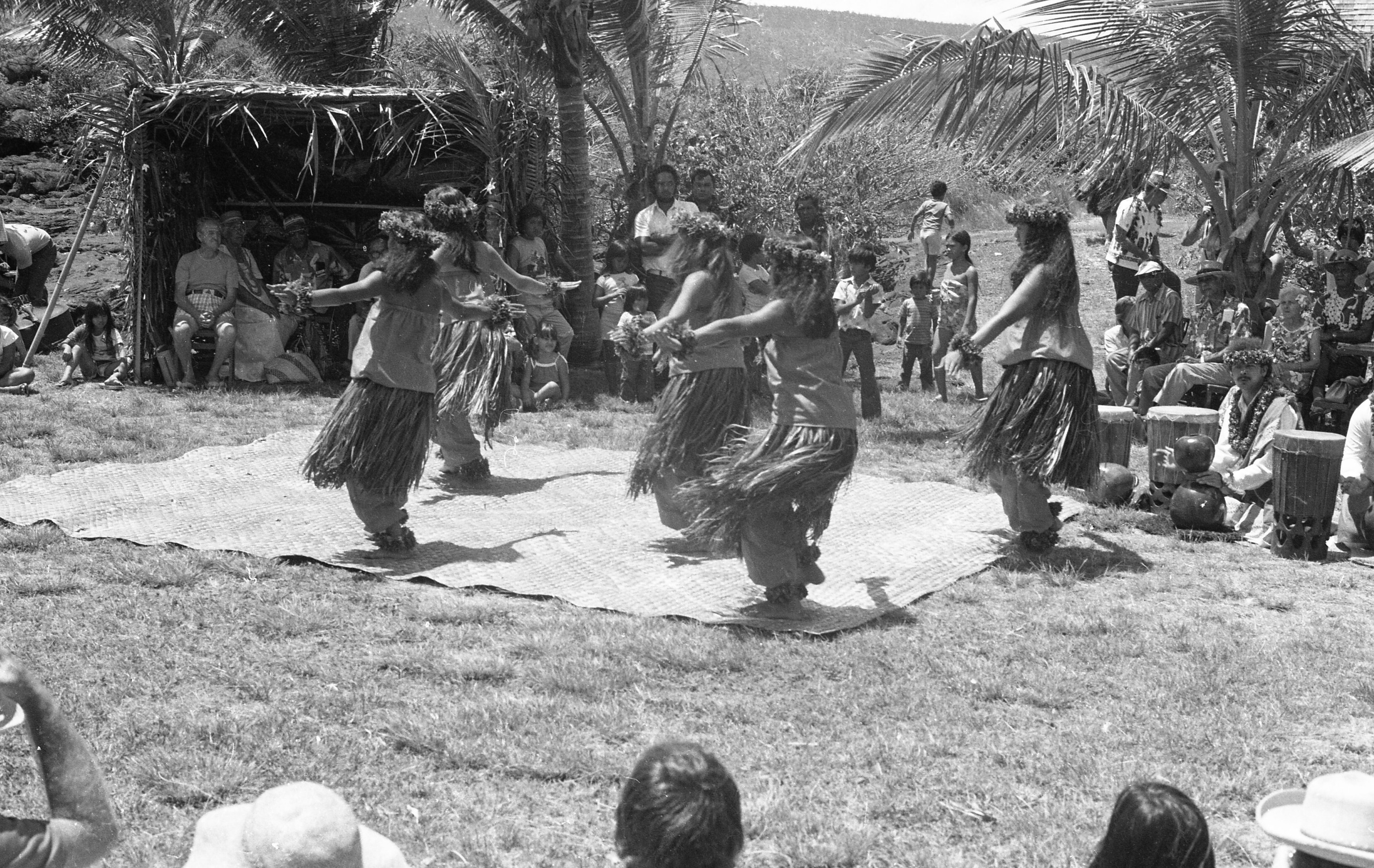 A black and white image of five women dancing on a woven mat during a cultural festival. They are dancing on a woven mat in two rows. The row in front closest to the camera has two women dancing and the back row has three. They are dressed similarly. Each woman is wearing a floral headband, lei, floral bracelets, one on each wrist, a tube top, a grass skirt with pants underneath, and a floral anklet on each of their feet They are facing towards the upper left corner of the image with their backs towards the camera. Their knees are bent slightly, and their arms are stretched out to their sides. In the foreground of the image the back of three people's heads is visible. In the background there are other people sitting on the ground, in chairs, and standing watching the performers. Some of the people are seen sitting inside of a booth constructed out of palm tree leaves towards the top left side of the image.