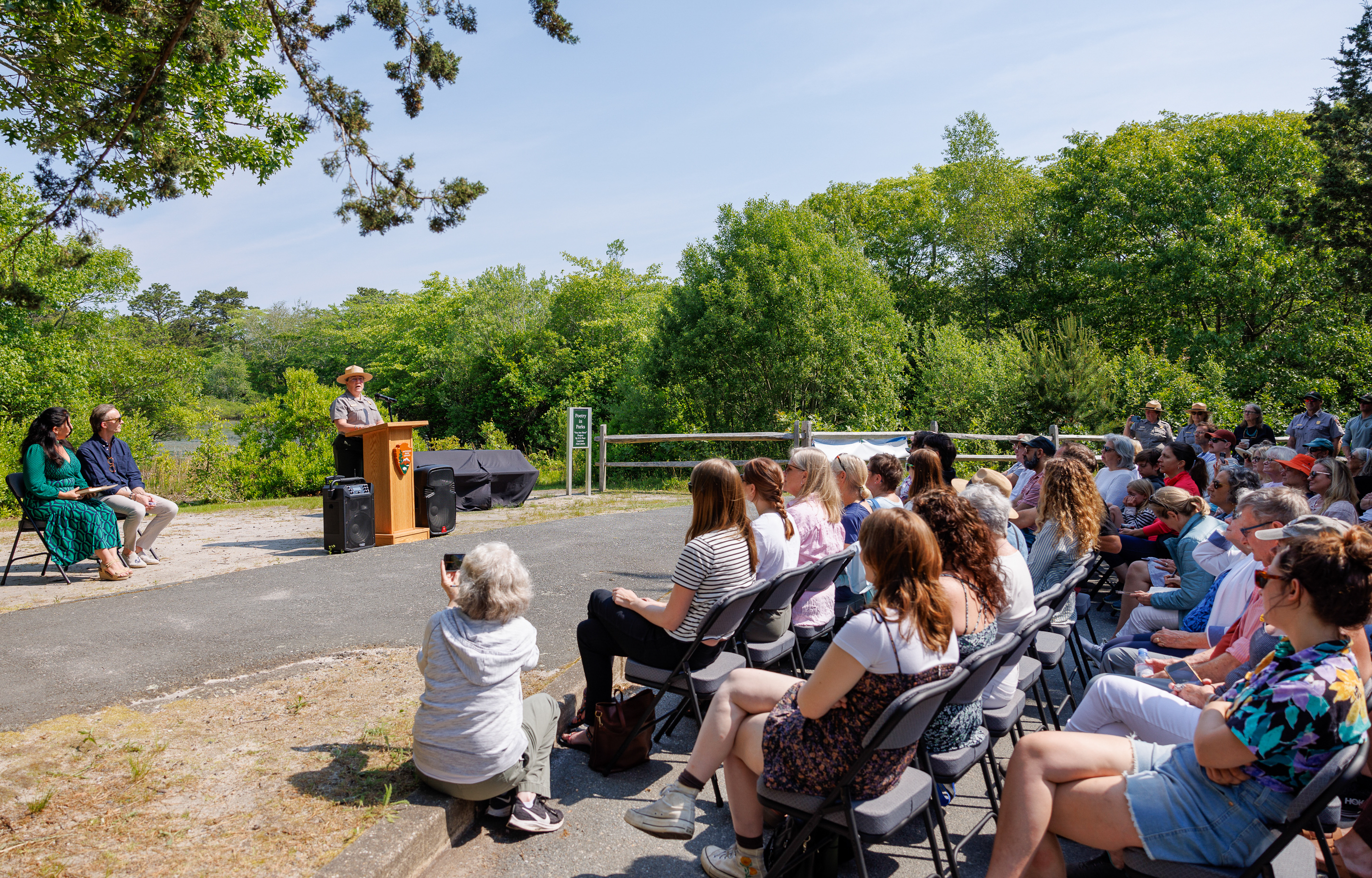 A person speaks at a podium in front of a seated audience at an outdoor installation unveiling.