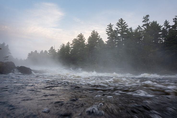 Fast moving white water is visible in the middle ground while dark water in the foreground is blurred from movement. Fogg rises from the river water and trees line the shoreline.