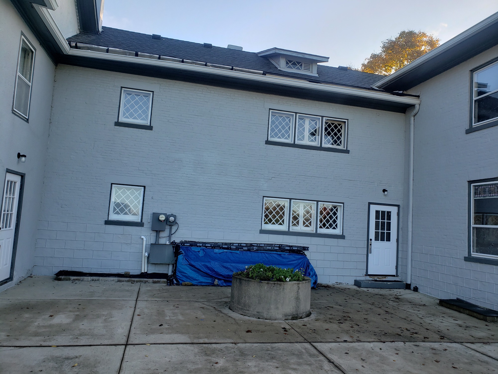 Two story grey brick home with multiple windows and white door above a step, blue tarp covering a section near grey box, and cement circular garden with green plants.