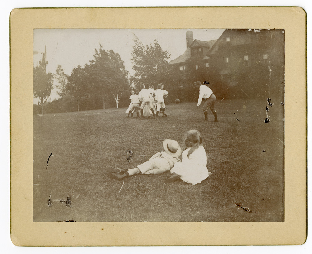 A group of children huddled with a football flying away from the huddle towards a man. A little boy in a straw hat and a little girl sit with their backs to the camera and look at the group playing football. A grand house and trees are in the background. 