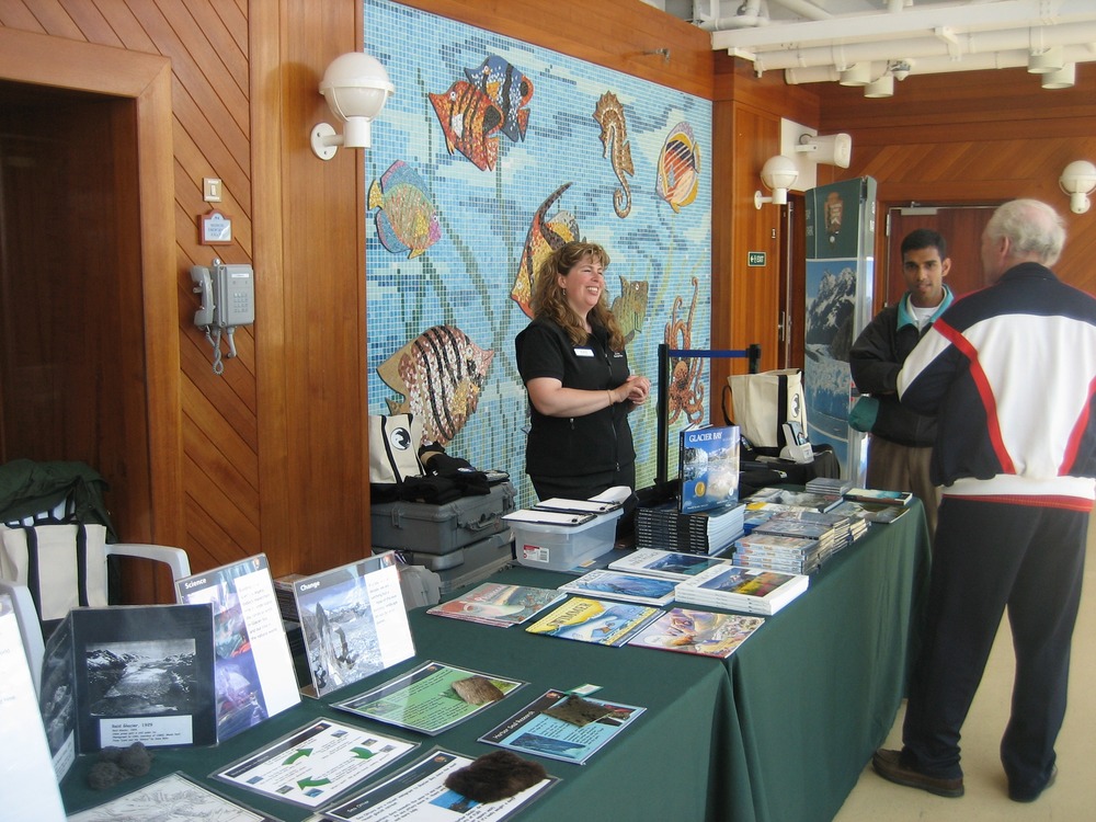 Woman dressed in black stands behind two tables covered in merchandise and educational materials. She is smiling as she interacts with visitors. 