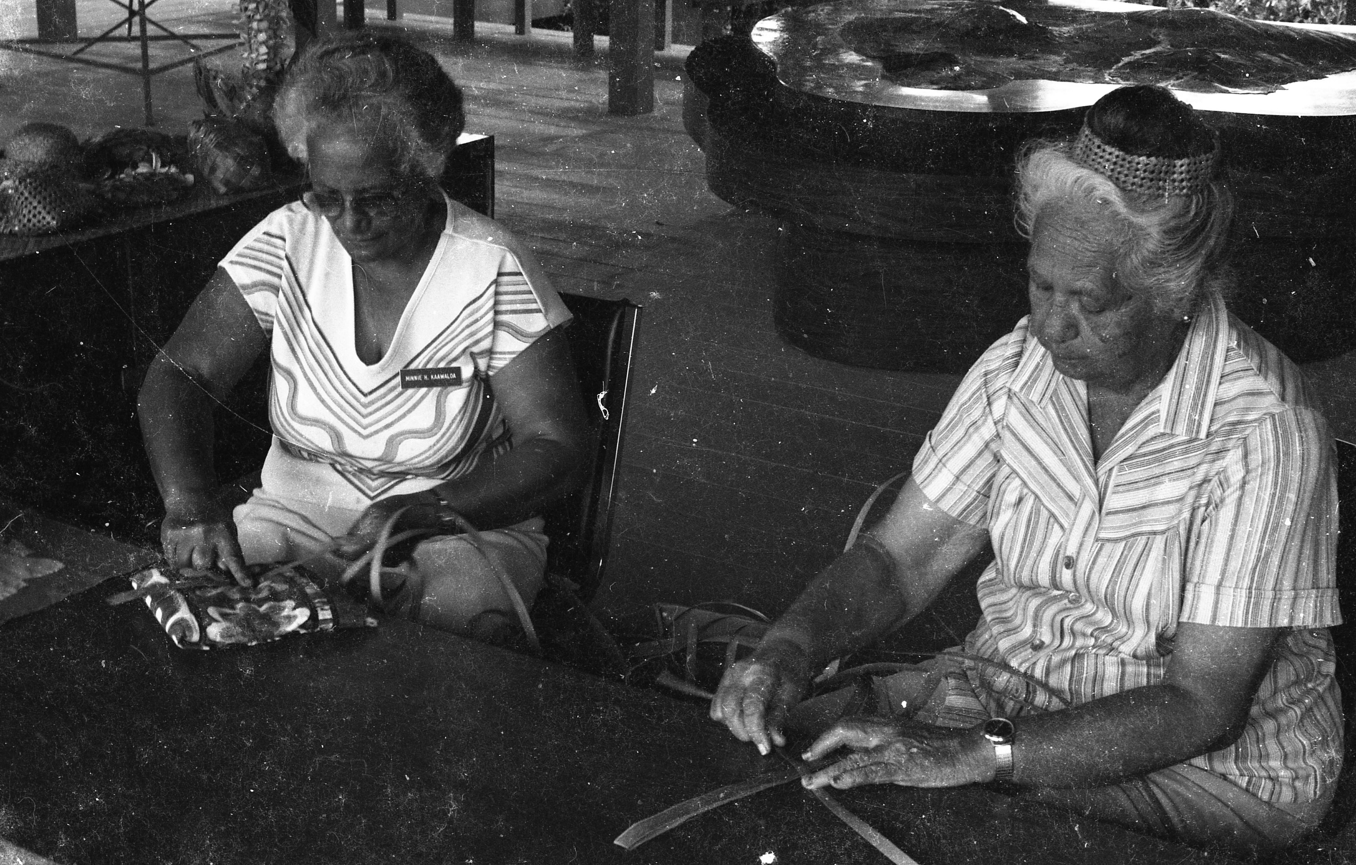 A black and white image of two women sitting down weaving at a table.  One of the women is located on the left side of the image. She is sitting on a chair. She has short hair and is wearing glasses and a short sleeve shirt. She has a name tag on the top left side of her shirt that reads, "Minnie H. Ka'awaloa." She is holding a tool in her right hand and a strip of a palm leaf in her left. She has the leaf propped up on a small pillow on a large table. She is pressing the tool into the width of the palm tree leaf. In between this woman and the other woman there is a pile of palm tree leaves. On the right side of the image is the second woman who is also sitting on a chair. She has her hair up in a bun and is wearing earrings, a button-down shirt, and a watch on her left wrist. Her hands are holding onto a small woven square with long strips of palm leaves sticking out. In the background there is a wooden floor and a large wooden table with a raised map of Hawaiʻi on top.