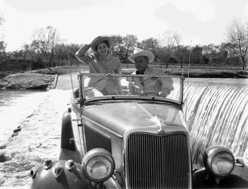 Both in cowboy hats, Senator and Mrs. Johnson drive a Ford Phaeton across a flooded low water crossing.