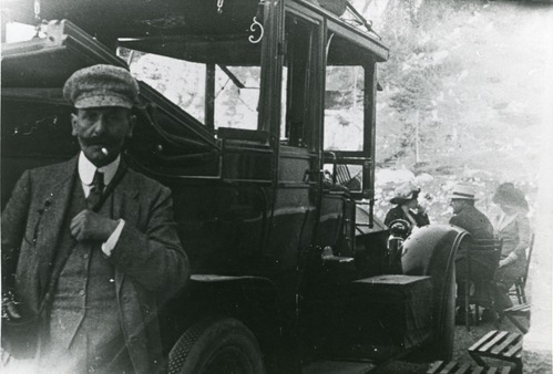 Man in a three-piece suit standing by a car. In the background are two women and a man sitting. Location, Pordoi Pass, Italy.