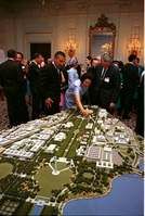 A crowd in business attire gathers in an elegant room of the White House, with three individuals leaning over an architectural model of the Washington DC Mall area with buildings, trees, streets, and waterways. 