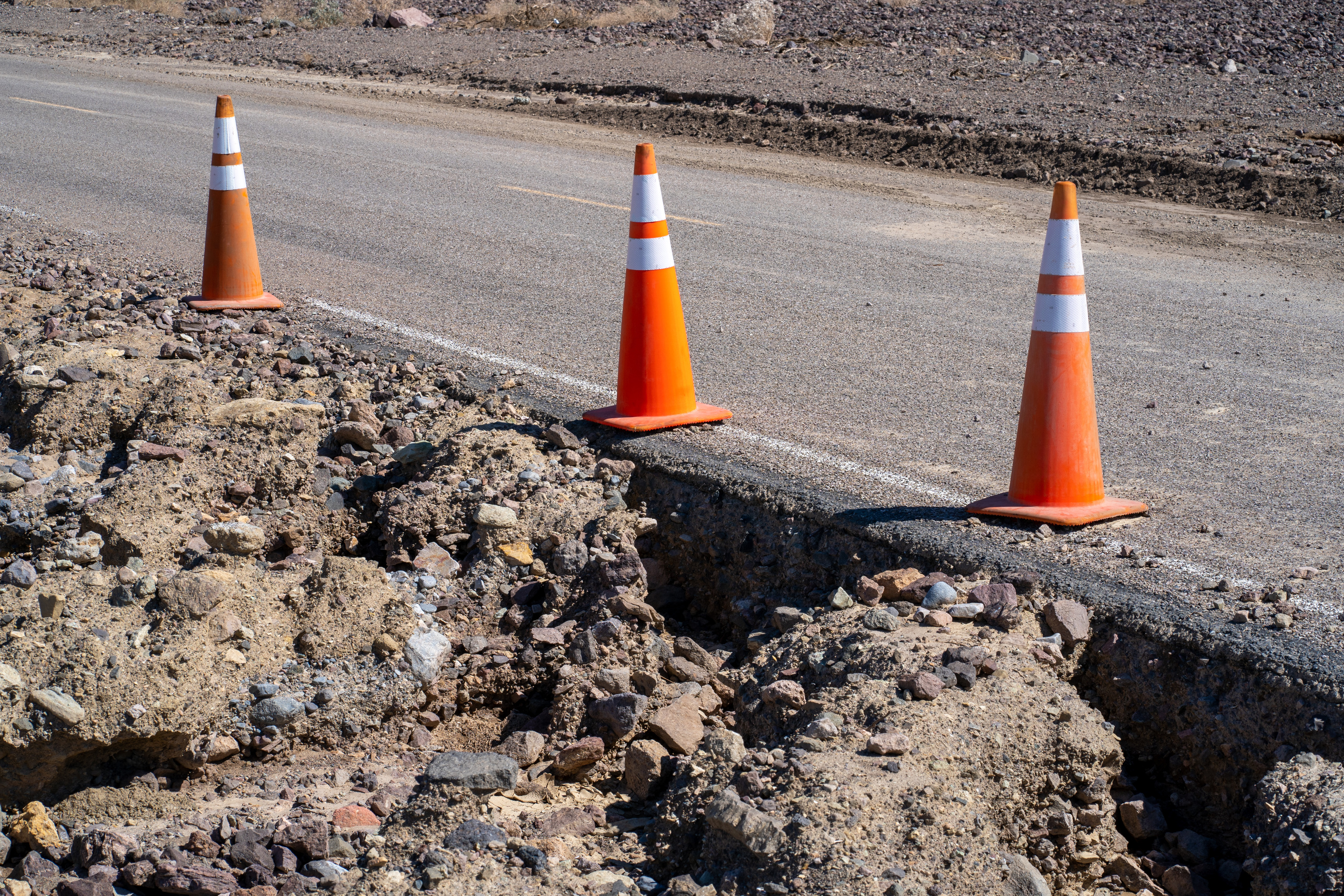Undercut road shoulder with debris including large rocks. Three orange cones sit on the asphalt road.