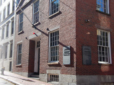 Photograph of a three story brick building at a streetcorner. First story on main street has a centered doorway flanked by two rectangular windows. The upper story has three equally spaced windows. Out of the photograph is a third story under a pediment. On both sides of the building's corner, a sign is visible.