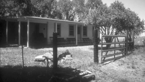 A one-story white ranch house is surrounded by trees, fencing of mixed materials, and a firepit in the lawn.