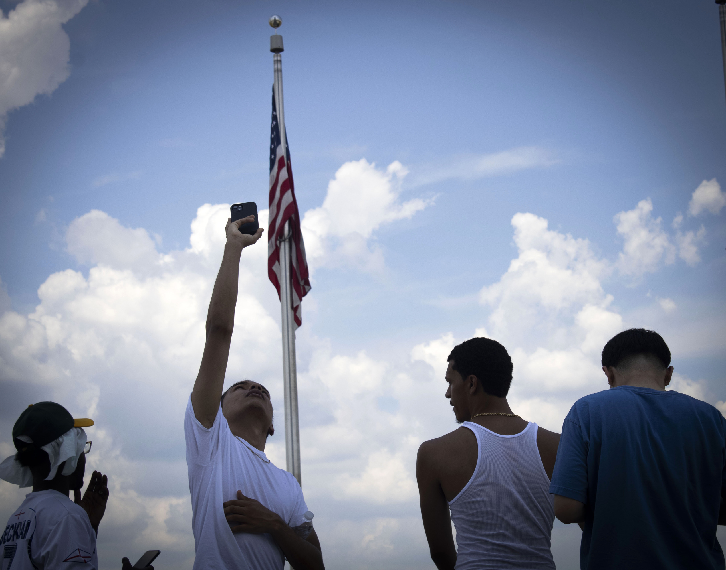 A group of young men stand in front of an American Flag at the Washington Monument. One of the men is holding is cell phone up towards the sky. 