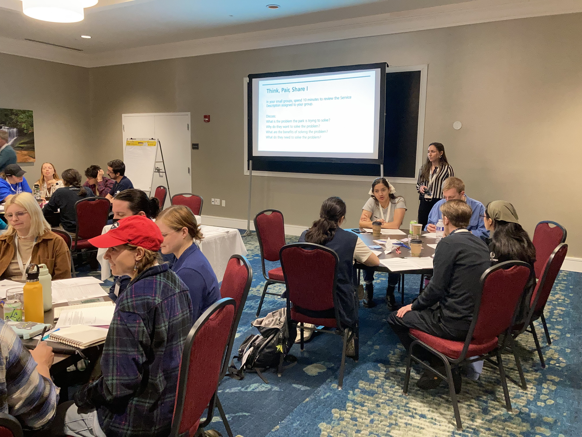 Several groups of Community Volunteer Ambassadors (CVAs) sit at round tables in a board room, a speaker stands next to a lit projection screen. 