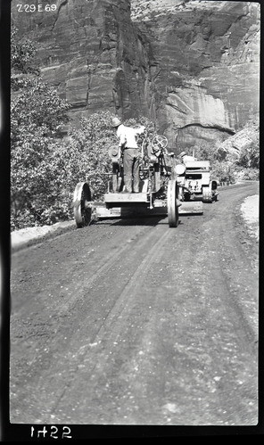 Worker driving a road grader following another worker on a caterpillar tractor on the floor of the valley road spreading paving materials. Labeled 'experimental road paving.'